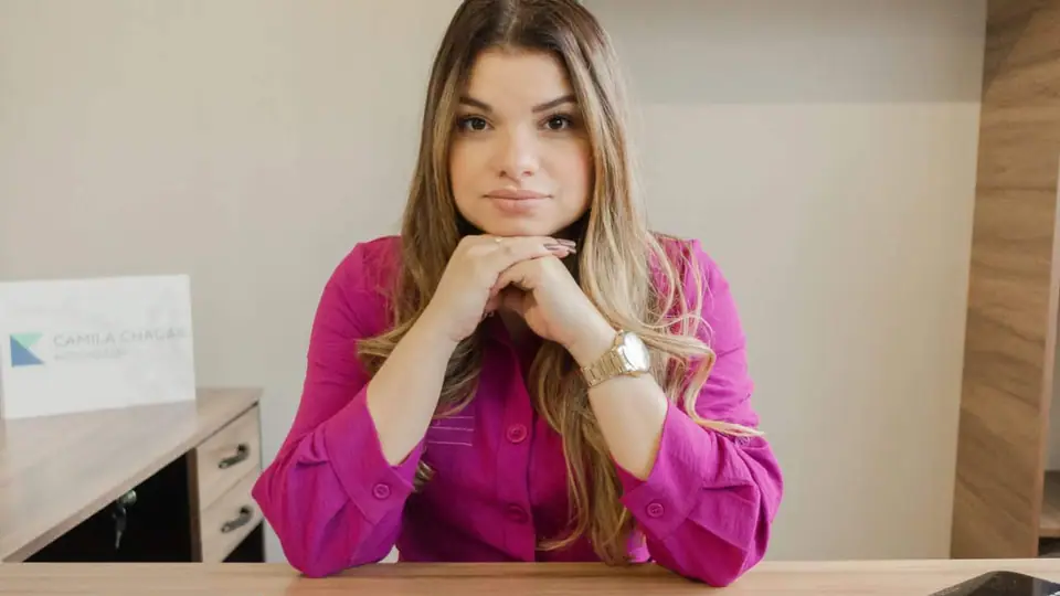 Dra. Camila Chagas à mesa do escritório, de camisa magenta, com as mãos apoiadas sob o queixo