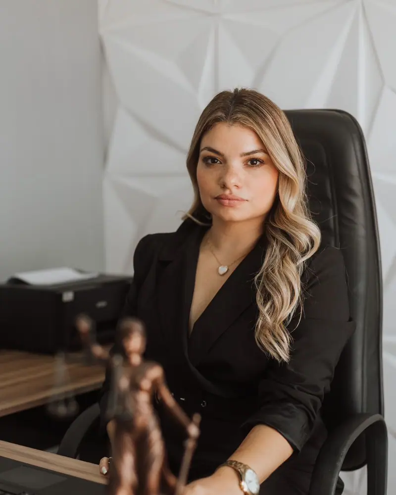 Dra. Camila Chagas sentada à mesa do escritório, de blazer preto, com a estatueta da Justiça em primeiro plano