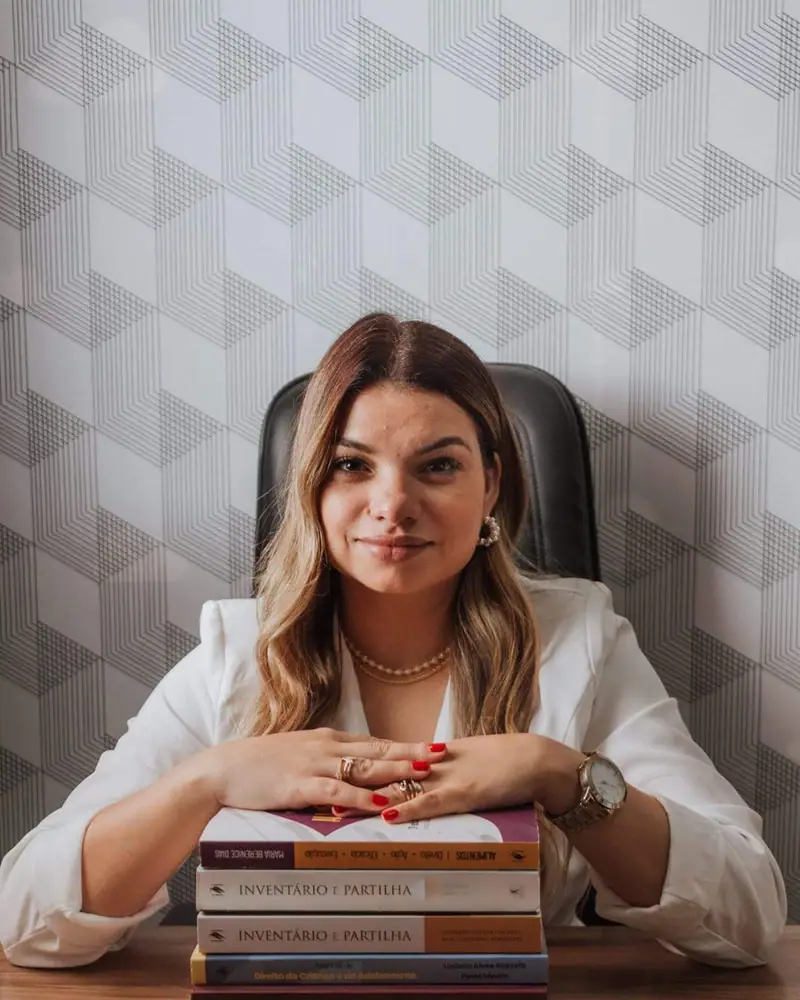 Dra. Camila Chagas à mesa, de blazer branco, com as mãos sobre uma pilha de livros de Direito